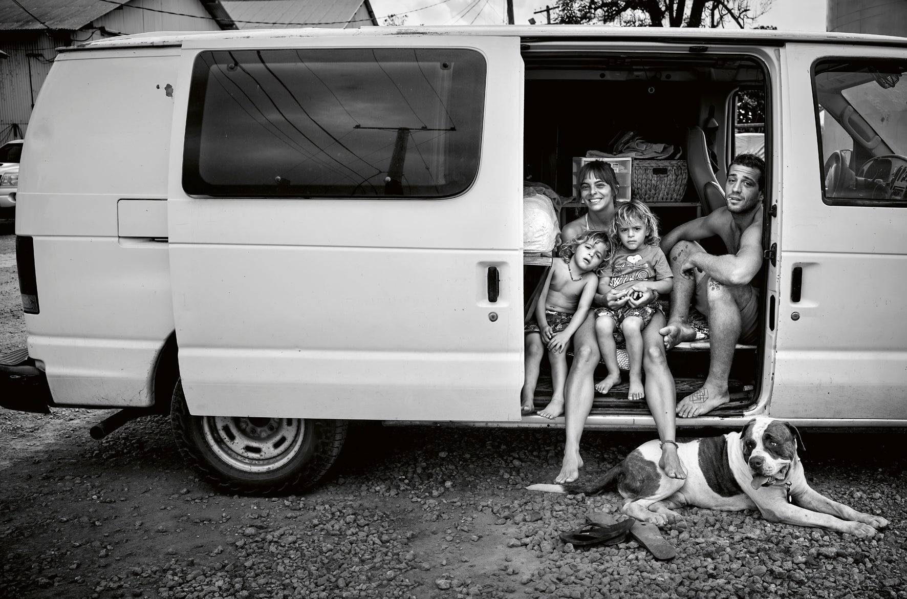Family with children and a dog inside a van on a gravel surface.