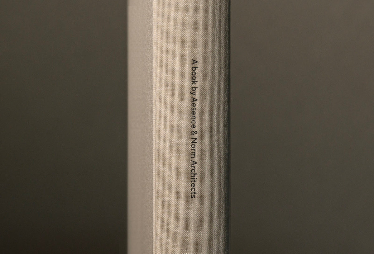 Close-up of a book spine with a textured beige linen cover and black text reading 'A book by Assense & Norm Architects' against a dark taupe background.