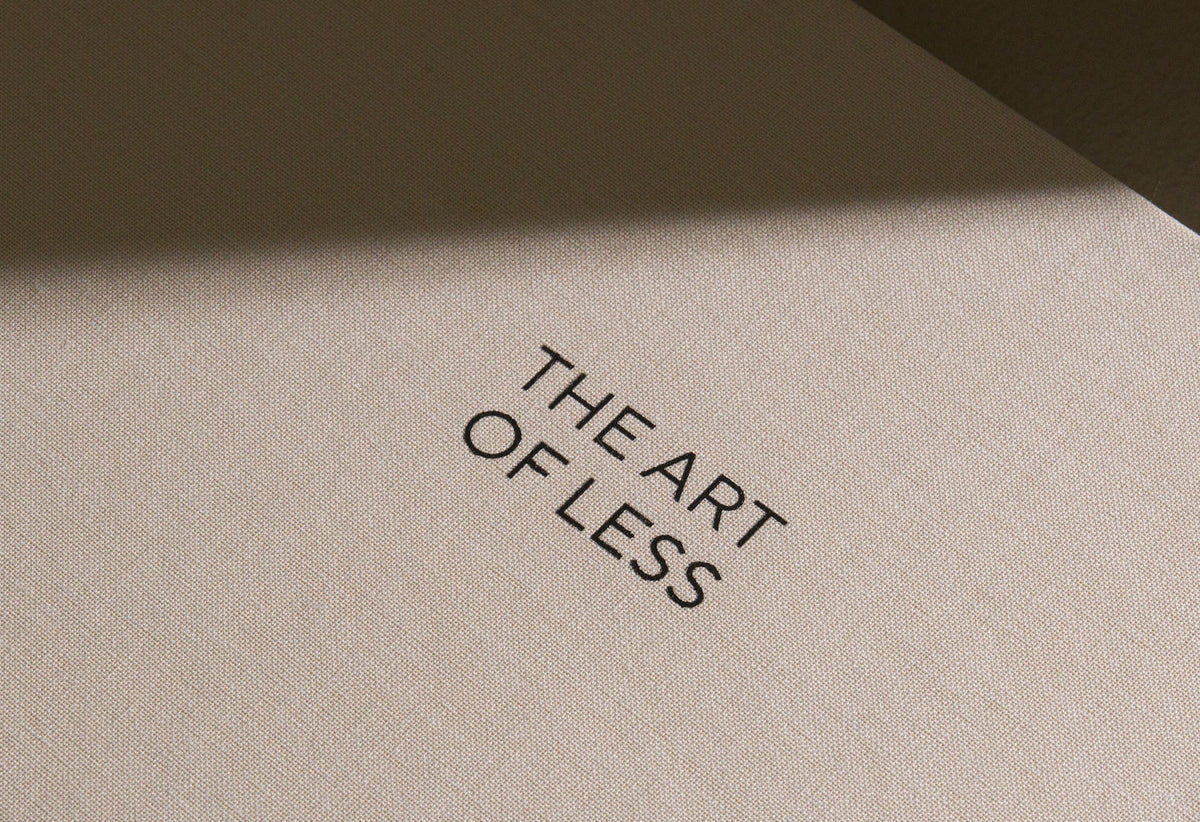 A close-up of a textured beige surface with the phrase 'THE ART OF LESS' printed in black serif text, lit by a soft diagonal shadow.