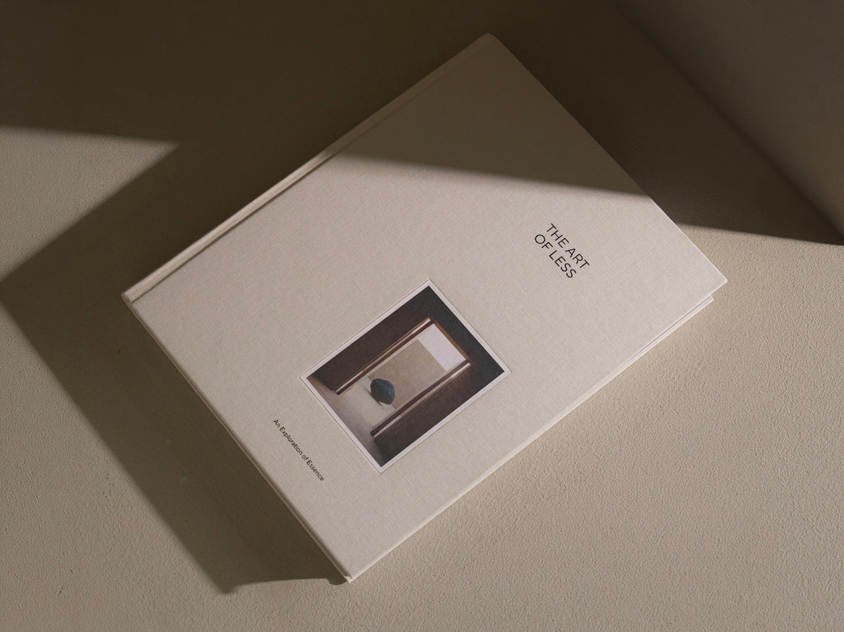 A cream-colored book titled 'The Art of Less' lies on a matching textured surface under dramatic, warm directional sunlight casting a sharp shadow.