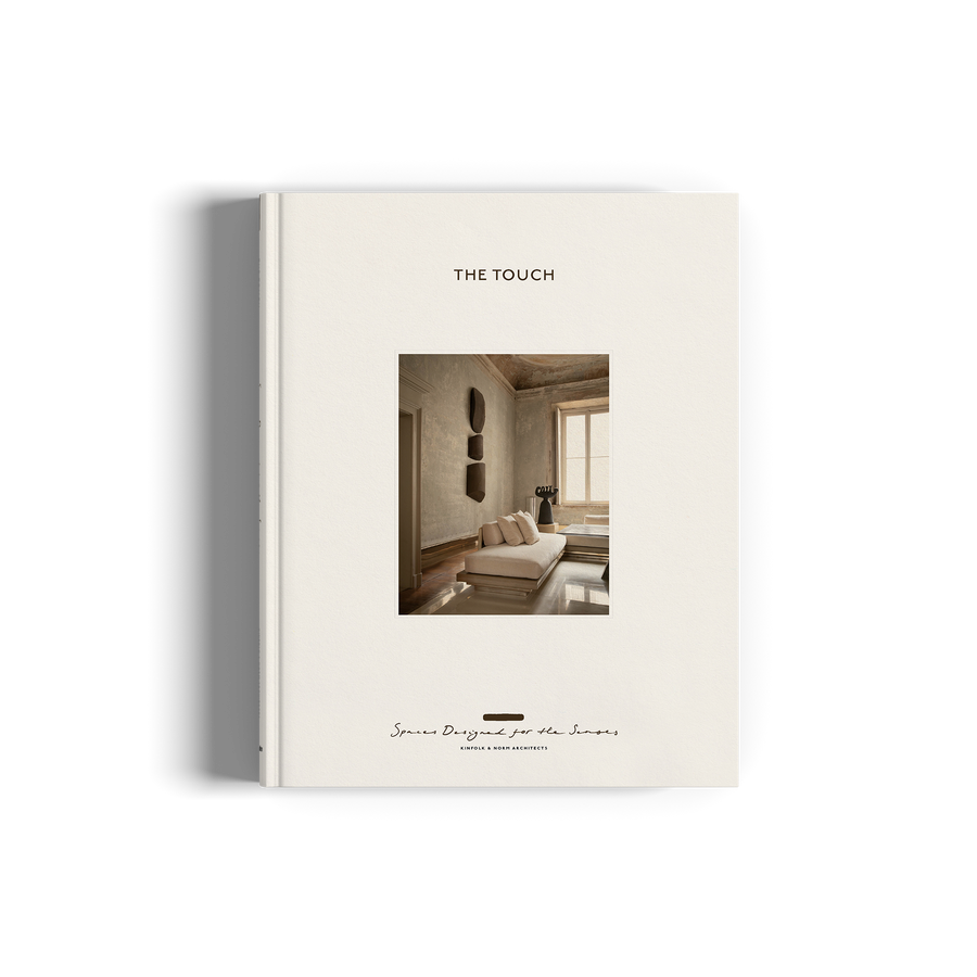 The image shows the cover of a book titled 'The Touch' with a white background. The cover features a minimalist design, depicting a serene indoor scene with a person seated in a chair, surrounded by simple, clean lines and a few decorative objects.
