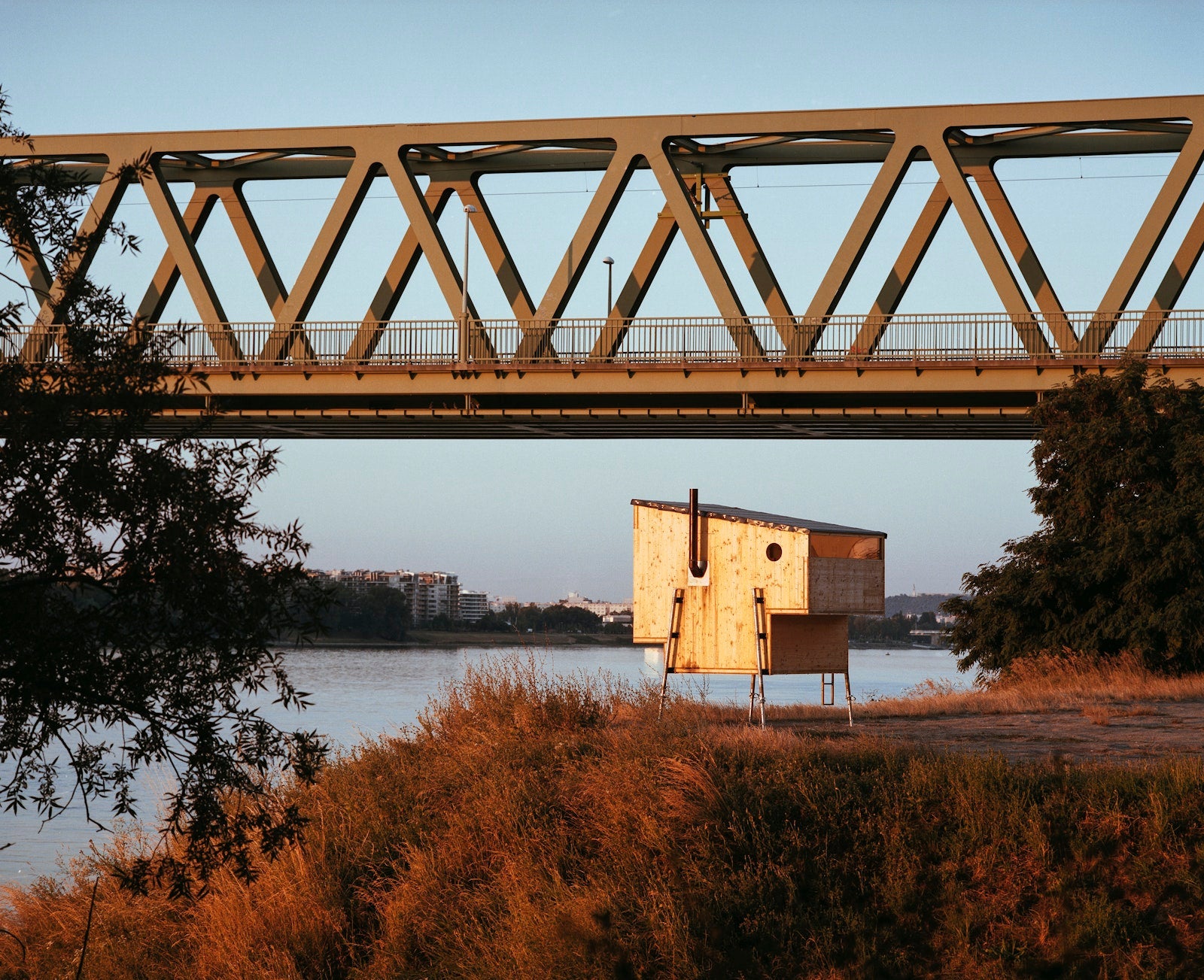 Wooden cabin by a river with a bridge in the background