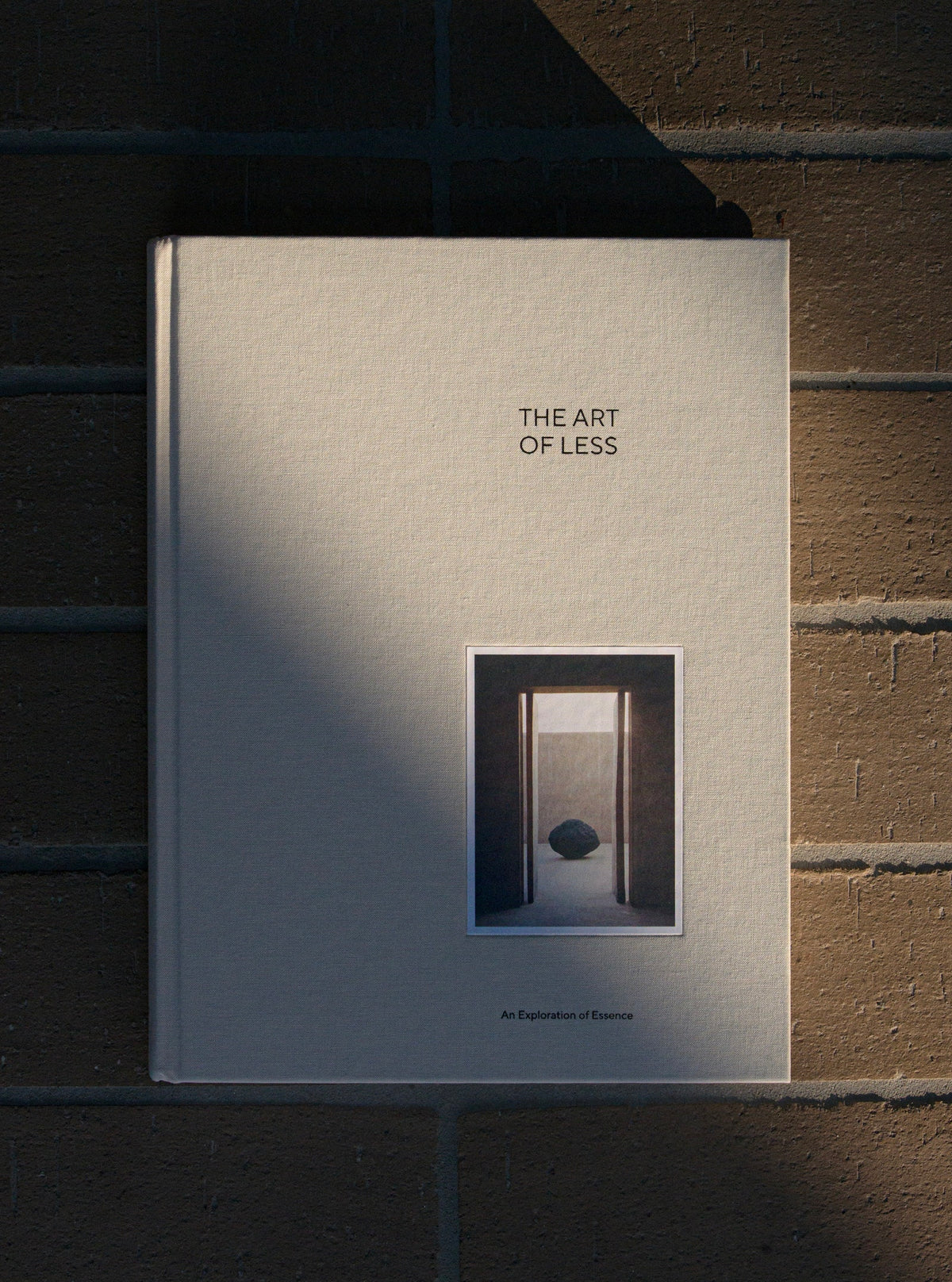 A hardcover art book titled 'THE ART OF LESS' resting against a brown brick wall, partially shaded by a strong diagonal shadow.