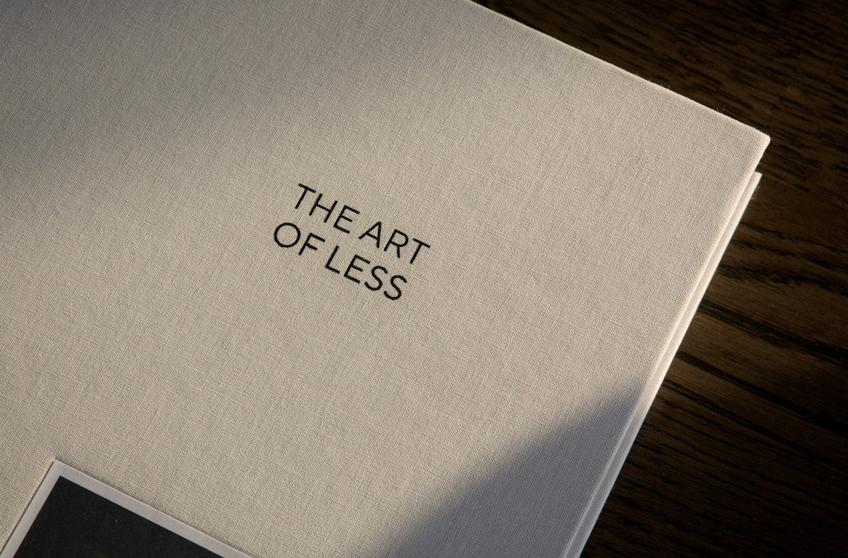A close-up of a white textured book or album cover with the title 'THE ART OF LESS' printed in black serif text, resting on a dark wood surface.