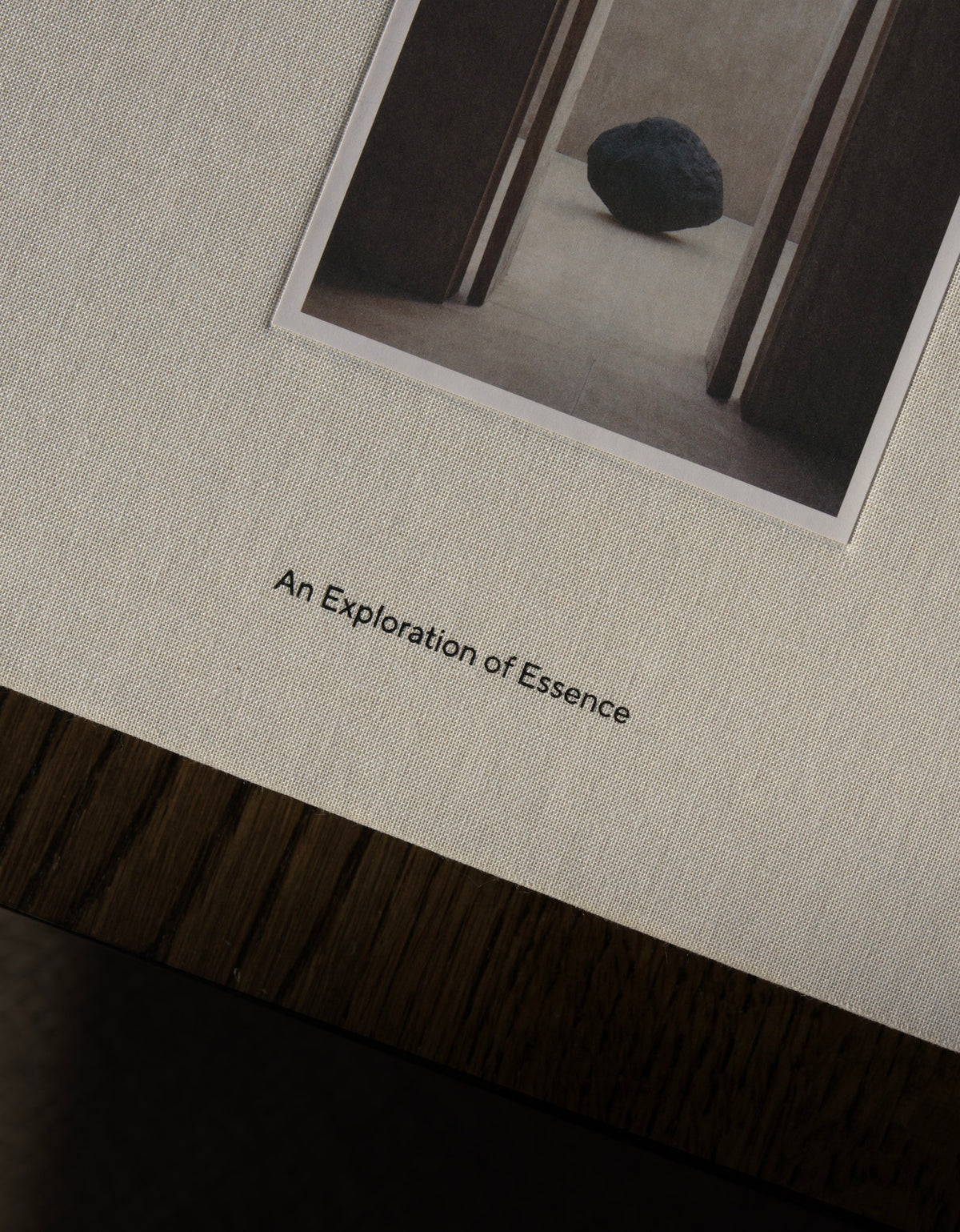 A close-up of a textured cream-colored book or art print cover, featuring a small black-and-white photograph of a dark stone in a doorway and the title 'An Exploration of Essence' printed below.