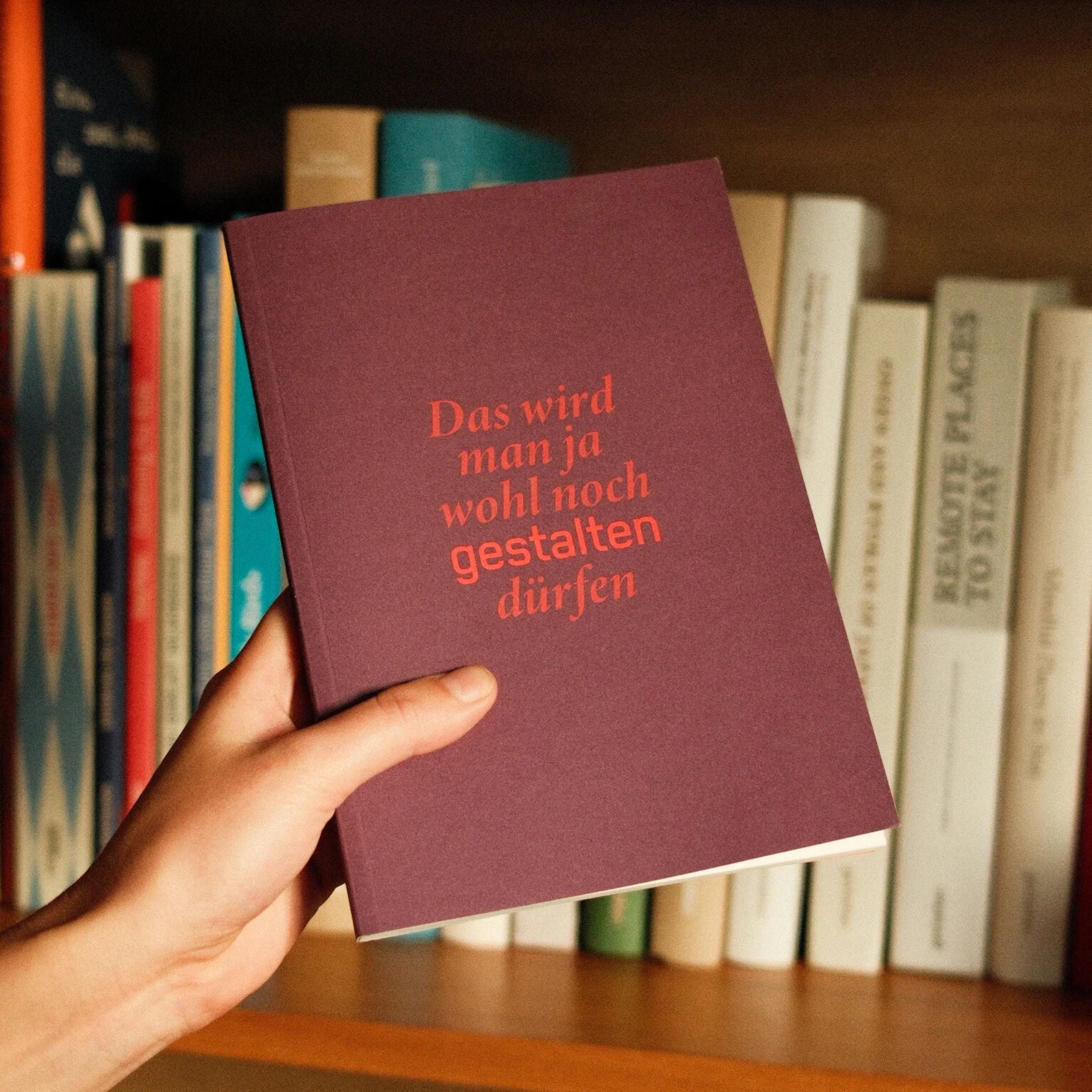 Hand holding a maroon book with red text in front of a bookshelf