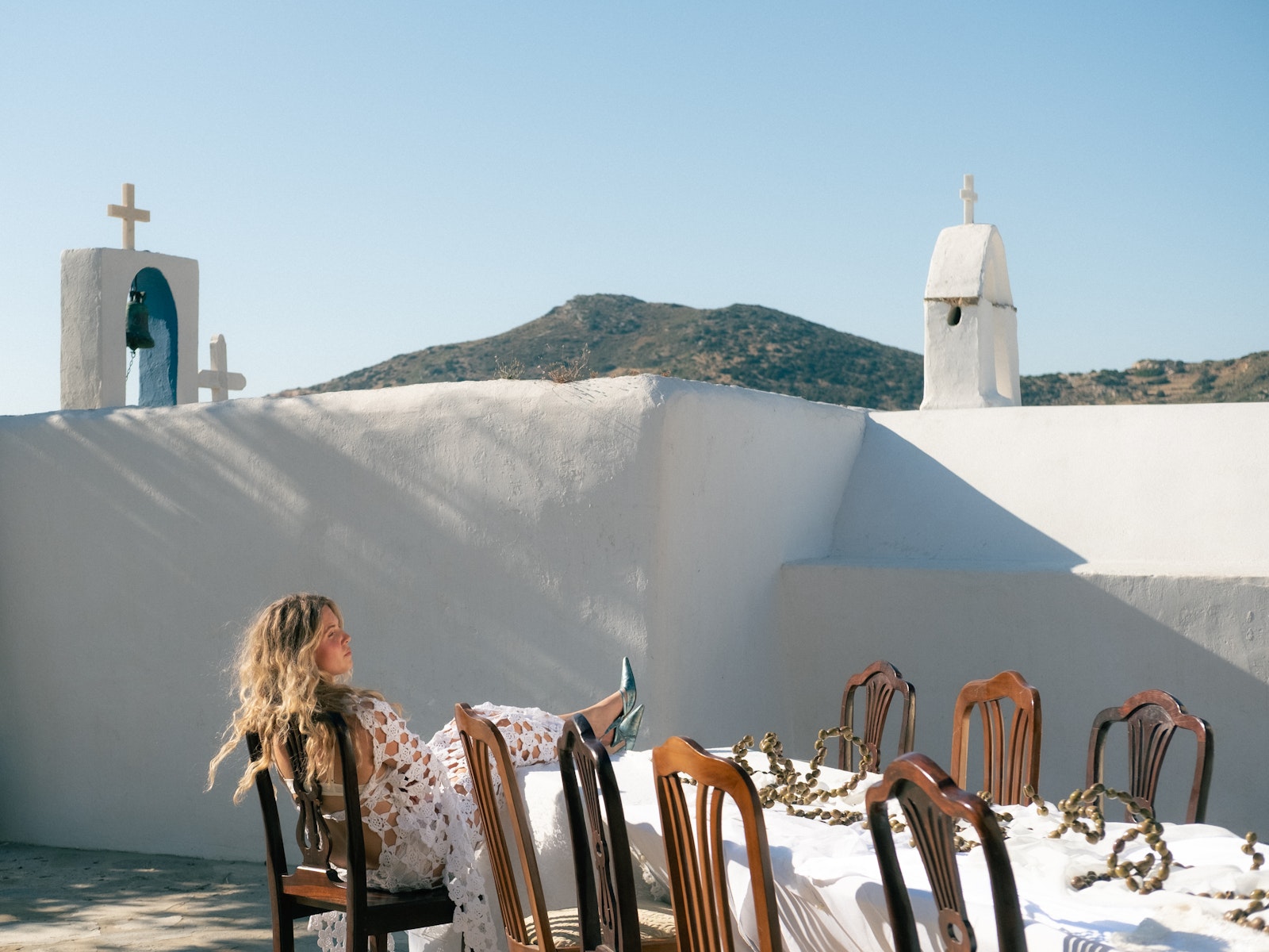 Woman sitting at a table with chairs on a rooftop with white walls and bell towers in the background.