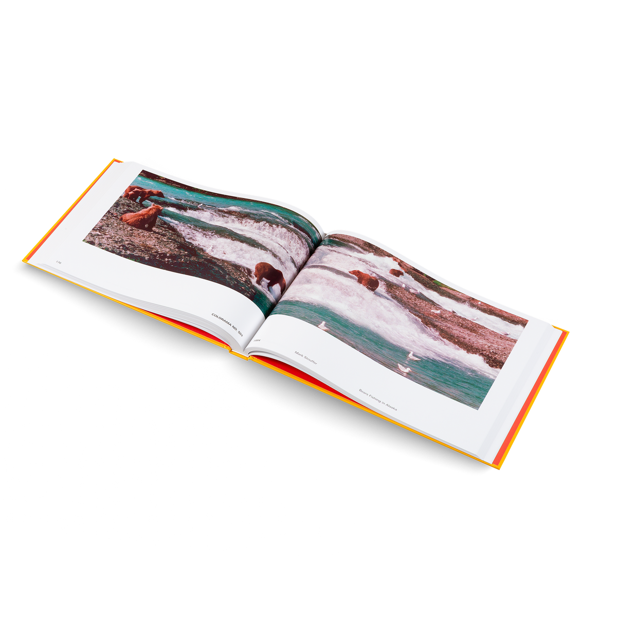 An open hardcover book photographed on a clean white background, angled slightly to the left. The pages show a colorful landscape photograph with flowing turquoise water, white surf, and dark rocky shoreline.