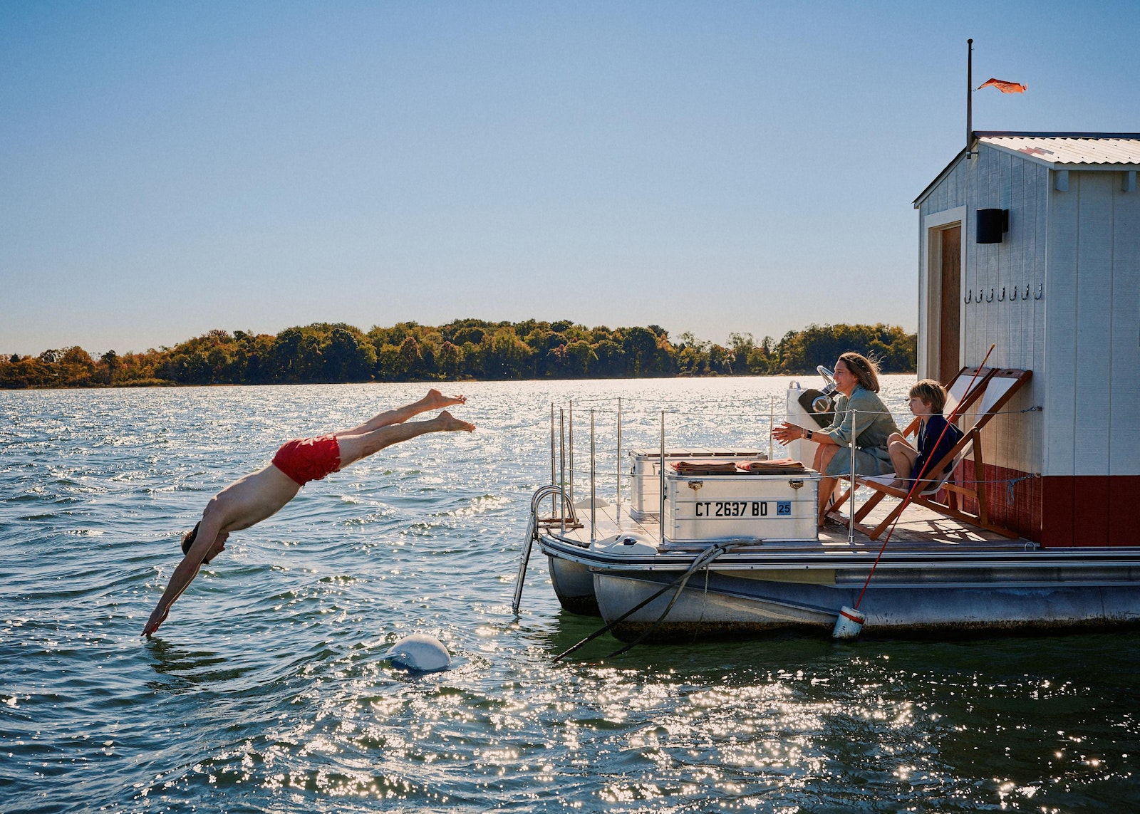 Person diving into water from a dock with a houseboat in the background