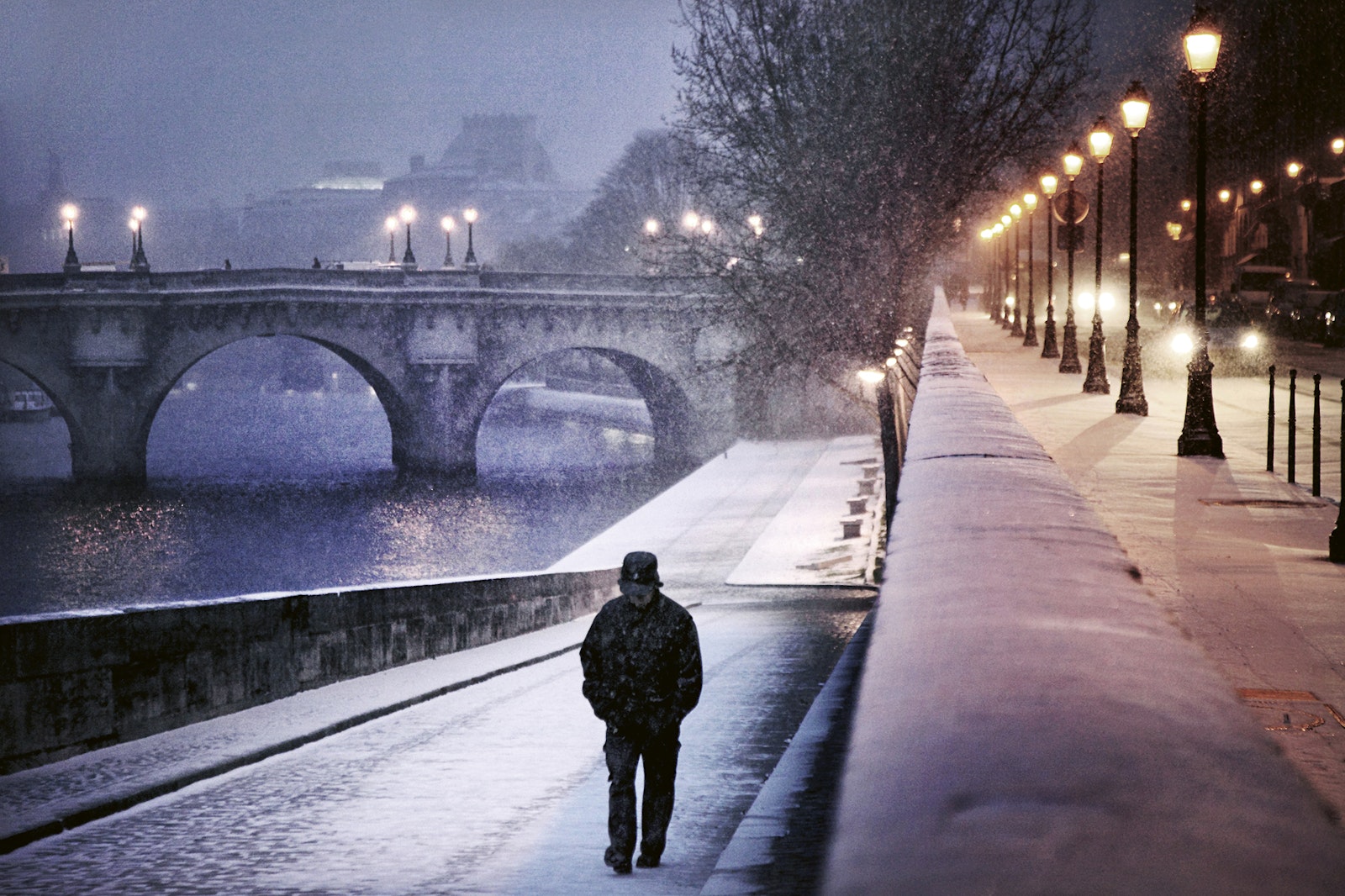 Person walking on a snow-covered street next to a river with a bridge in the background
