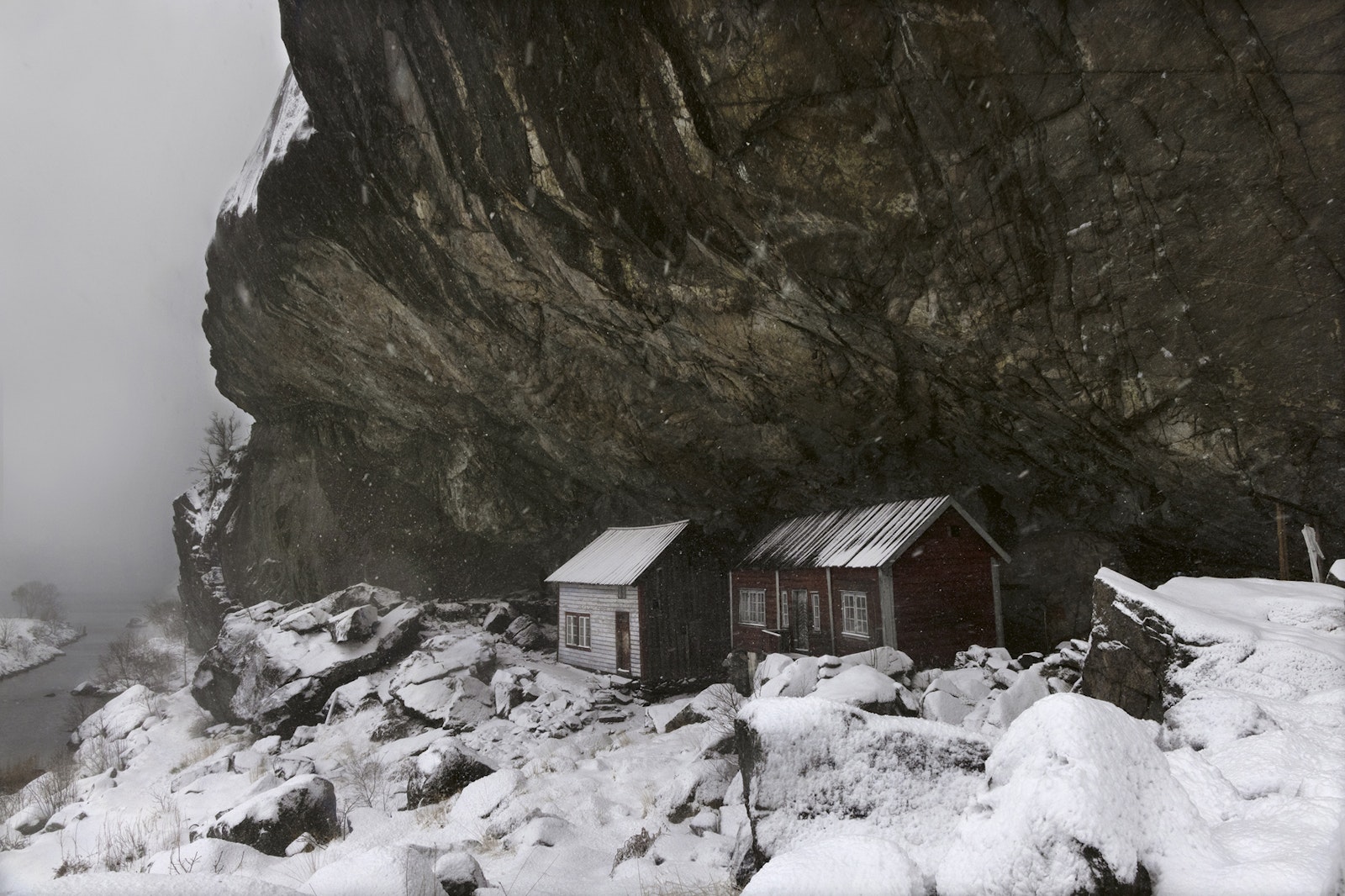 Two small cabins nestled against a rocky cliffside with snow-covered ground.