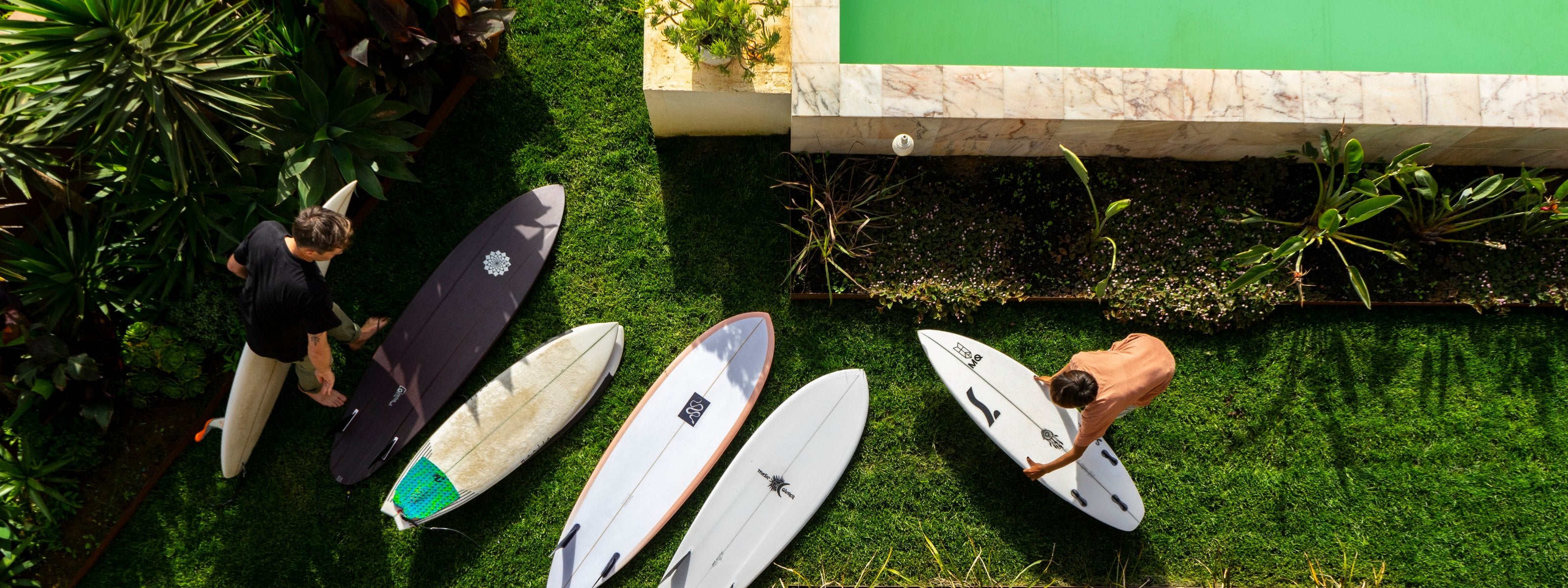 Two people with surfboards on a grassy area next to a pool.