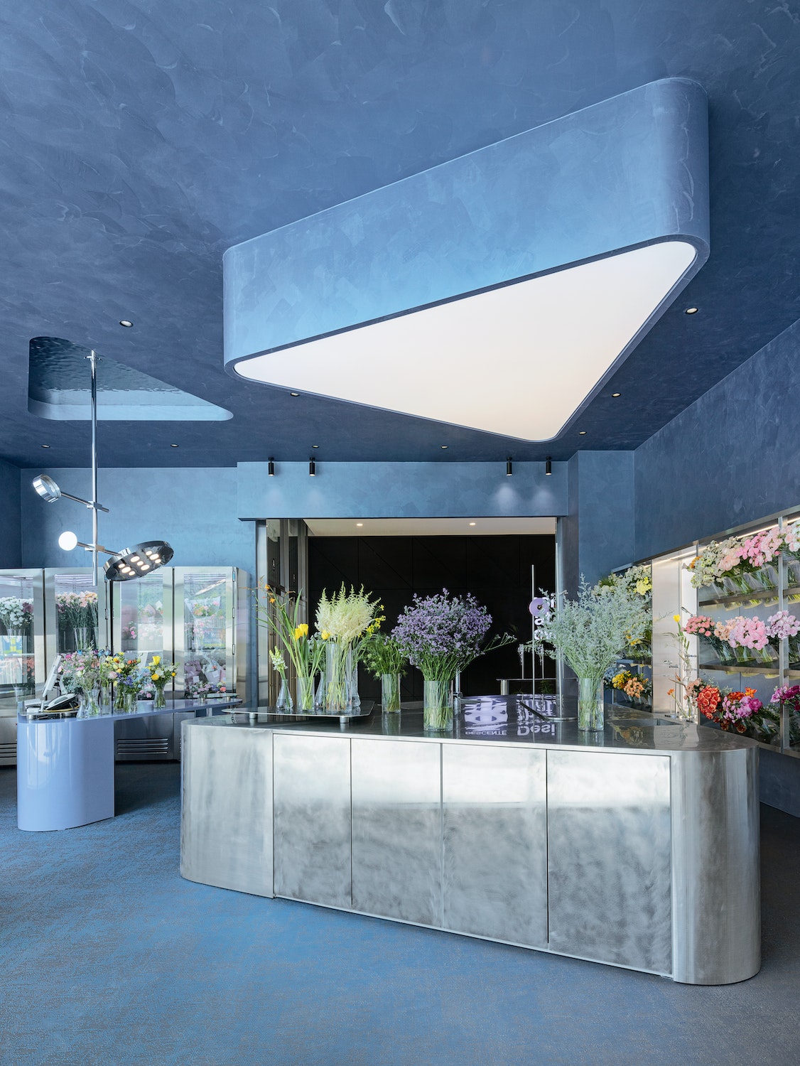 Irgendwo zwischen Labor und Spaceshuttle bewegt sich die Ästhetik von o’flower dem ersten Store des Onlineblumenhändlers Okkot. Das in Seoul ansässige Architekturbüro Plainoddity entwarf das Interieur als unaufdringliche Kulisse, die der Blumenpracht den Vortritt lässt. Vom frischen Himmelblau der Wände hebt sich die Metalltheke ab, deren Form vom Oberlicht zitiert wird. Daneben versetzt der große Tisch mit acht Stühlen in Erstaunen – in einem Blumenladen?